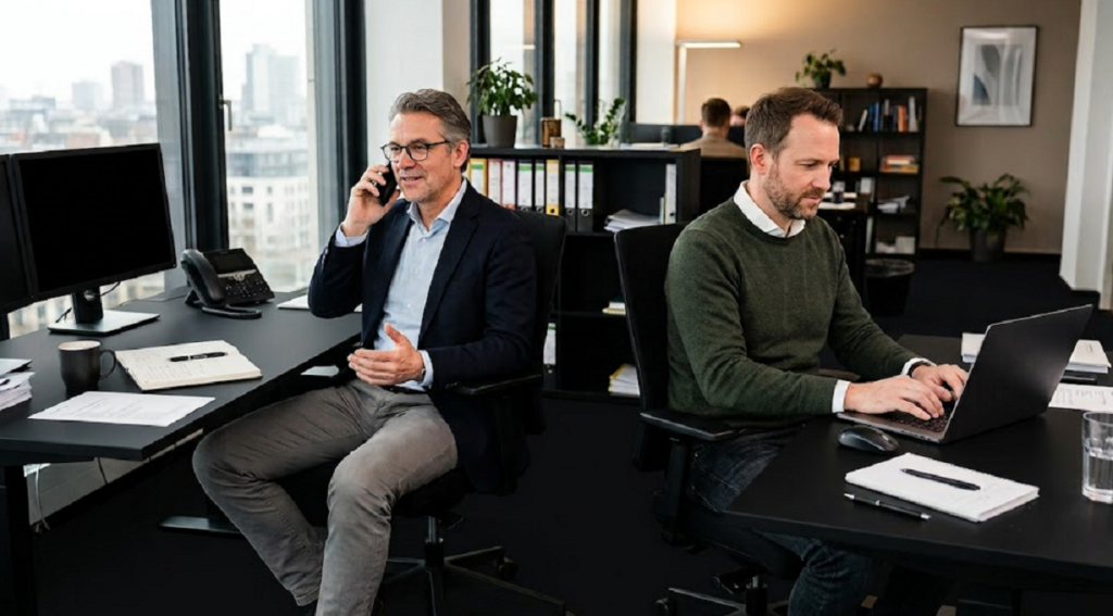 Zwei Mitarbeiter arbeiten in einem modernen Büro: Ein Mann telefoniert, während ein Kollege an einem Laptop sitzt. Helle Büroräume mit Schreibtischen, Regalen und großen Fenstern.